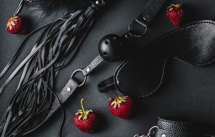 A stylish flat lay of leather BDSM accessories with fresh strawberries on a dark background.